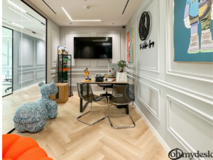 A stylish and modern office space with a minimalist design, featuring white paneled walls, a large flat-screen TV, and colorful artwork. The room is furnished with a small desk, clear acrylic chairs, and a blue patterned decorative piece resembling a stuffed animal.